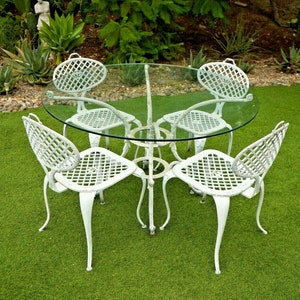 Pode incluir: Um conjunto de p&aacute;tio de ferro fundido branco com uma mesa redonda de vidro e quatro cadeiras. As cadeiras t&ecirc;m um design de treli&ccedil;a e est&atilde;o dispostas em torno da mesa sobre uma superf&iacute;cie de grama artificial verde.