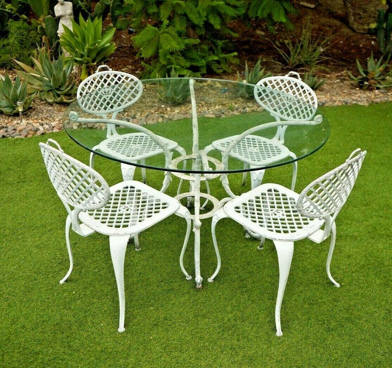 Pode incluir: Um conjunto de p&aacute;tio de ferro fundido branco com uma mesa redonda de vidro e quatro cadeiras. As cadeiras t&ecirc;m um design de treli&ccedil;a e est&atilde;o dispostas em torno da mesa sobre uma superf&iacute;cie de grama artificial verde.