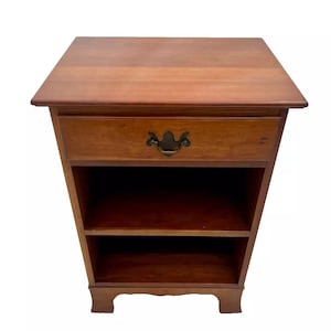 May include: A wooden end table with a single drawer and two shelves. The drawer has a brass pull with a decorative design. The table has a dark wood finish and sits on four legs.