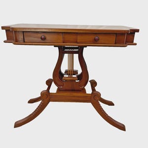 May include: A wooden side table with a lyre-shaped support and two small drawers. The table is made of a warm-toned wood and features decorative carvings on the legs and apron. The drawers have round knobs.