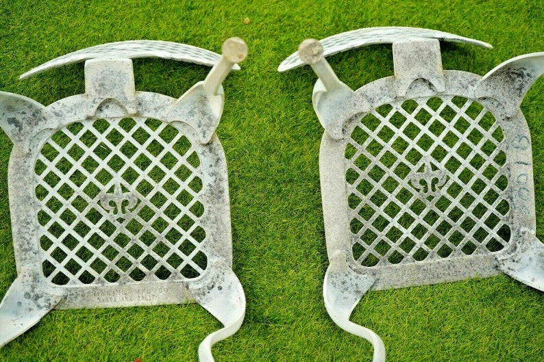 Pode incluir: Duas cadeiras de metal branco com um padr&atilde;o de grelha em forma de diamante no assento e no encosto. As cadeiras t&ecirc;m um design de flor-de-lis no centro do assento.