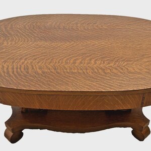 May include: An oval coffee table constructed from medium-brown wood featuring a prominent tiger oak grain pattern on the tabletop surface. The table is supported by a central pedestal base with four curved feet extending outward at the bottom.