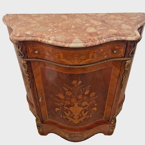 May include: A semi-circular wooden cabinet featuring a mottled pink and brown marble top. The front displays intricate floral marquetry, a single drawer with two small knobs, and a central door with gold-toned metal decorative accents.