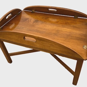 May include: A wooden folding tray table with a warm brown finish. The tray has curved edges with built-in handles and hinged side panels. The legs are angled for stability. The tray is designed for serving or displaying items.