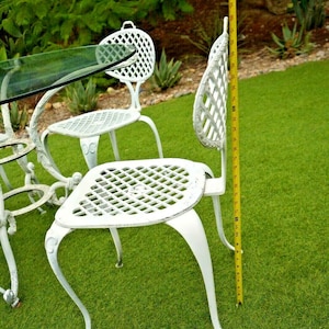 Pode incluir: Conjunto de m&oacute;veis de jardim de ferro fundido branco com uma mesa de vidro e quatro cadeiras. As cadeiras t&ecirc;m um design de treli&ccedil;a e medem 91 cm de altura.