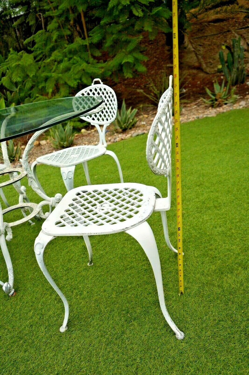Pode incluir: Conjunto de m&oacute;veis de jardim de ferro fundido branco com uma mesa de vidro e quatro cadeiras. As cadeiras t&ecirc;m um design de treli&ccedil;a e medem 91 cm de altura.