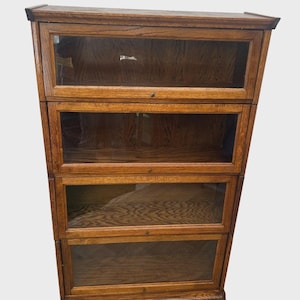 May include: A four-tiered wooden bookcase with glass-paned doors. The bookcase is made of a light brown wood with visible grain. Each section has a small, round knob for opening. The bookcase is empty.