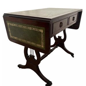 May include: A dark wood antique desk with a green leather top and two drawers. The desk has ornate legs and a decorative green panel with gold trim. The desk is a classic piece of furniture.
