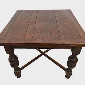 May include: A square wooden table with a dark brown finish and a visible wood grain pattern on the top surface. The table features four thick, turned legs with a rounded bulbous design and a cross-shaped stretcher connecting the legs at the base.