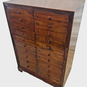 May include: A tall wooden cabinet featuring two vertical columns of drawers. Each drawer is equipped with a small round knob and is labeled with a letter of the alphabet, starting with A through M on the left and N through W on the right side.
