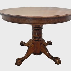 May include: A round wooden table with a medium brown finish and a visible wood grain pattern. The table features a central pedestal base with a turned column design and four legs that terminate in carved claw and ball feet at the base.