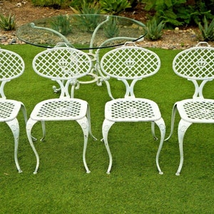 Pode incluir: Um conjunto de quatro cadeiras de ferro fundido branco com um design de treli&ccedil;a decorativo e uma mesa com tampo de vidro. As cadeiras e a mesa est&atilde;o dispostas em um gramado verde.