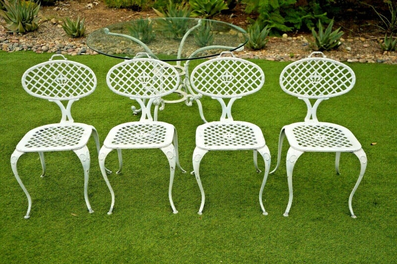 Pode incluir: Um conjunto de quatro cadeiras de ferro fundido branco com um design de treli&ccedil;a decorativo e uma mesa com tampo de vidro. As cadeiras e a mesa est&atilde;o dispostas em um gramado verde.