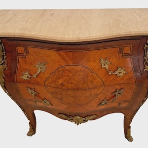 May include: A curved wooden chest of drawers with a light-colored marble top. The chest features ornate brass hardware and a decorative wood grain pattern. The drawers have a curved front and are supported by curved legs.