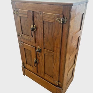 May include: A vintage wooden cabinet with a warm, medium-brown finish. The cabinet features two sets of double doors with brass hardware, including hinges and handles. The design is rectangular with a slightly angled top.