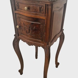 May include: A wooden bedside table featuring a light gray marble top, one drawer with a small brass knob, and a single cabinet door with carved floral details. The piece stands on four curved cabriole legs with intricate wood carvings at the top.