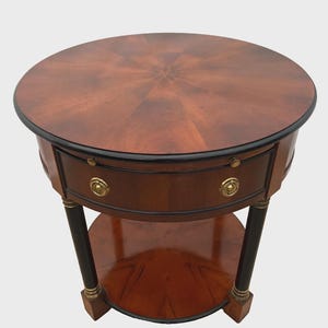 May include: A round, wooden side table with a dark brown finish and a black trim. The table features a drawer with a brass knob and a lower shelf. The legs are black with gold accents. The table is approximately 24 inches in diameter.