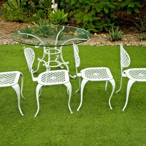 Pode incluir: Conjunto de m&oacute;veis de jardim de metal branco com uma mesa de vidro e quatro cadeiras. As cadeiras t&ecirc;m um design de treli&ccedil;a e pernas curvas.