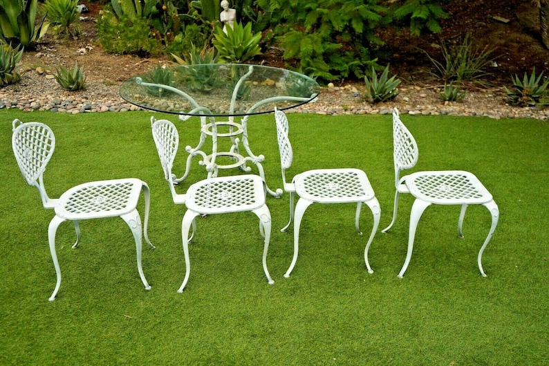 Pode incluir: Conjunto de m&oacute;veis de jardim de metal branco com uma mesa de vidro e quatro cadeiras. As cadeiras t&ecirc;m um design de treli&ccedil;a e pernas curvas.