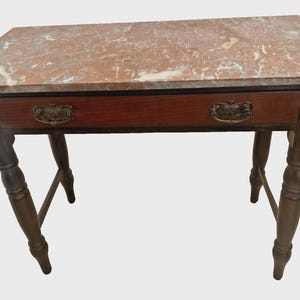 May include: A rectangular wooden table featuring a polished reddish-brown marble top with white veining. The table includes a single horizontal drawer with two ornate brass pull handles and four turned legs connected by a lower support frame.