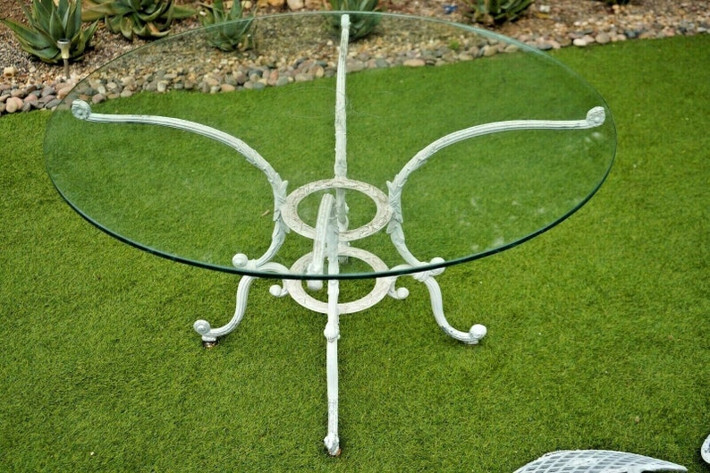 Pode incluir: Mesa de p&aacute;tio de ferro forjado branco com tampo redondo de vidro. A mesa tem um design decorativo e ornamentado.