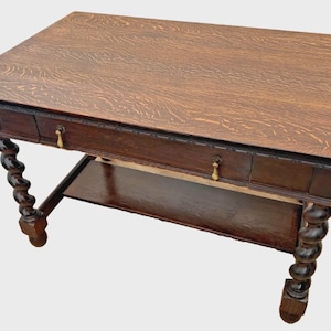 May include: A rectangular wooden desk with a prominent grain pattern on the top surface. The desk features a single long drawer with two brass drop handles and four spiraled, twisted legs connected by a lower rectangular shelf base.