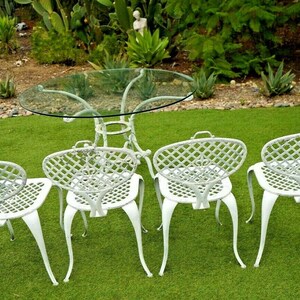 Pode incluir: Um conjunto de jantar de exterior em metal branco com uma mesa redonda de vidro e quatro cadeiras. As cadeiras t&ecirc;m um design de treli&ccedil;a e est&atilde;o dispostas em torno da mesa.