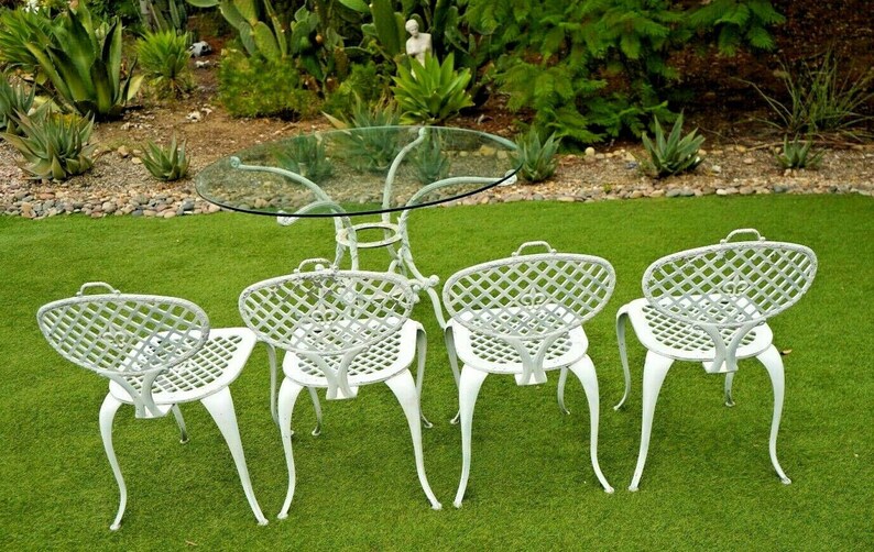 Pode incluir: Um conjunto de jantar de exterior em metal branco com uma mesa redonda de vidro e quatro cadeiras. As cadeiras t&ecirc;m um design de treli&ccedil;a e est&atilde;o dispostas em torno da mesa.