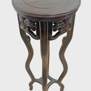 May include: A dark brown wooden pedestal table with a circular top and a smooth, polished finish. The table features four curved, slender legs that flare outward and connect at a central base, with intricate carved detailing under the top rim.