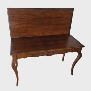 May include: A dark brown wooden desk with a raised back panel. The desk features a rectangular tabletop with a decorative floral carving along the front edge. The legs are curved, and the wood grain is visible throughout.