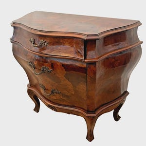 May include: A vintage, two-drawer chest of drawers with a curved front and ornate brass handles. The wood has a rich, dark brown finish with visible grain patterns. The chest rests on four curved legs, showcasing a classic antique design.