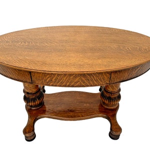 May include: A vintage oval-shaped wooden table with a lower shelf and turned legs. The table is made of oak wood and has a rich brown finish.