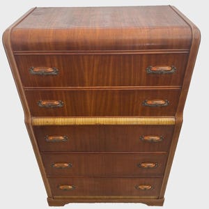 May include: A tall wooden chest of drawers featuring a rich brown finish with a smooth, rounded top edge. It includes five horizontal drawers, each equipped with two decorative metal handles. A textured, light-colored horizontal band accents the front.