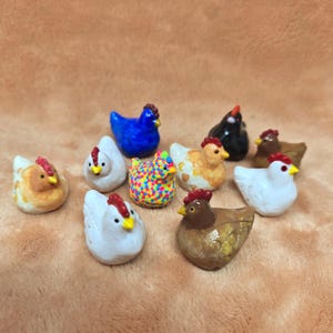 May include: A collection of small, decorative ceramic chickens in various colors. The chickens have red combs and yellow beaks. Some are white, brown, blue, or black, while one is covered in colorful dots. These figurines are likely for home decor.