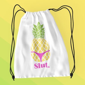 May include: White drawstring bag featuring a yellow pineapple wearing a pink bikini. The pineapple has green leaves and the word "Slut" in pink. Black drawstrings are visible.