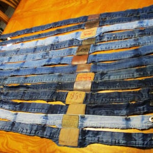 May include: A collection of ten blue denim jean waistbands arranged in parallel rows. Each waistband features a leather or fabric brand patch at the center and a metal button closure at the end. The denim shades range from light to dark blue.