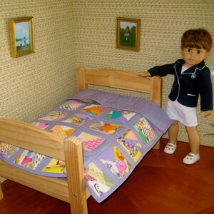 May include: A doll-sized bedroom scene featuring a wooden bed with a patchwork quilt in shades of purple, pink, and yellow. A doll in a navy blazer and white shorts stands beside the bed. Two small framed pictures hang on the textured wall.