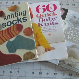 May include: A stack of knitting books, including "Knitting Socks" and "60 Quick Baby Knits." The "Knitting Socks" book features sock illustrations and yarn balls. The books are on a cutting mat.