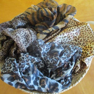 May include: A collection of lightweight scarves featuring various animal print patterns, including leopard and tiger designs in shades of brown, black, gray, and tan, contained within a round woven basket with two white handles.