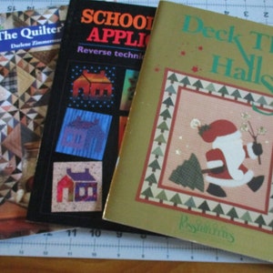 May include: Three quilting pattern books with colorful covers. The first book is titled "The Quilter" and features a quilt with a geometric pattern. The second book is titled "Schoolhouse Applique" and features a colorful house design. The third book is titled "Deck the Halls" and features a green and red Santa Claus design.