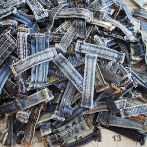May include: A large collection of blue denim belt loops in various shades of indigo, ranging from light to dark wash. Each individual loop features reinforced stitching in gold, white, or blue thread, showcasing frayed edges and worn fabric textures.