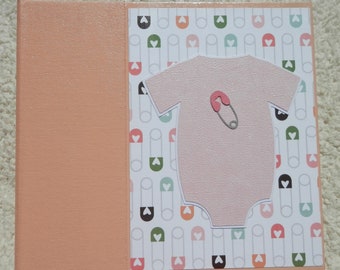 May include: A pink baby onesie with a safety pin on the front, against a background of white safety pins with pink, green, and orange hearts.