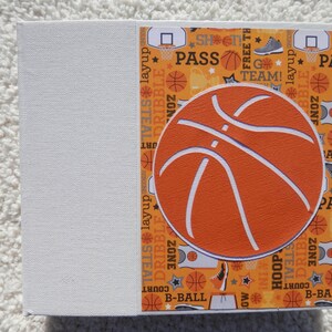 Puede incluir: Una caja de regalo cuadrada con tem&aacute;tica de baloncesto. La caja presenta un gran gr&aacute;fico de baloncesto naranja sobre un fondo estampado con palabras e im&aacute;genes relacionadas con el baloncesto. El lado izquierdo de la caja es blanco liso.
