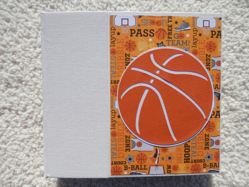 Puede incluir: Una caja de regalo cuadrada con tem&aacute;tica de baloncesto. La caja presenta un gran gr&aacute;fico de baloncesto naranja sobre un fondo estampado con palabras e im&aacute;genes relacionadas con el baloncesto. El lado izquierdo de la caja es blanco liso.