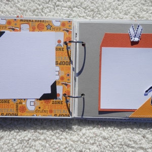Puede incluir: P&aacute;ginas abiertas de un &aacute;lbum de recortes con dise&ntilde;os de baloncesto. La p&aacute;gina izquierda tiene un marco de fotos blanco con esquinas negras, sobre un fondo naranja estampado con gr&aacute;ficos de baloncesto. La p&aacute;gina derecha presenta un fondo gris con un acento naranja y una silueta de jugador de baloncesto.