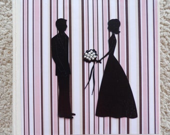May include: A white card with pink and brown stripes features a black silhouette of a couple. The bride is holding a bouquet of white flowers. The card is perfect for a wedding or engagement.