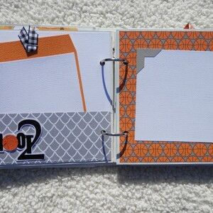 Puede incluir: P&aacute;ginas de &aacute;lbum de recortes abiertas con tem&aacute;tica de baloncesto. Incluye papel estampado naranja y gris, insertos para fotos y el texto "SHOOT 2". Una cinta a cuadros en blanco y negro a&ntilde;ade un toque decorativo.