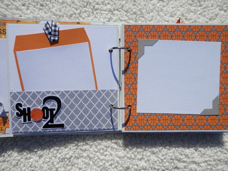 Puede incluir: P&aacute;ginas de &aacute;lbum de recortes abiertas con tem&aacute;tica de baloncesto. Incluye papel estampado naranja y gris, insertos para fotos y el texto "SHOOT 2". Una cinta a cuadros en blanco y negro a&ntilde;ade un toque decorativo.