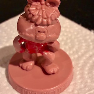 May include: A small pink plastic figurine of a pig character with a textured head, wearing a red shirt, standing on a circular base. The base features the embossed text LITE STUFF. The figure has a snout, ears, and a pose with one hand on its hip.