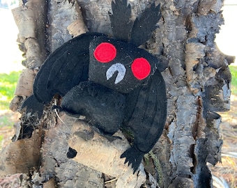 May include: A black felt plush toy shaped like a moth with large red eyes. The moth is perched on a tree trunk.